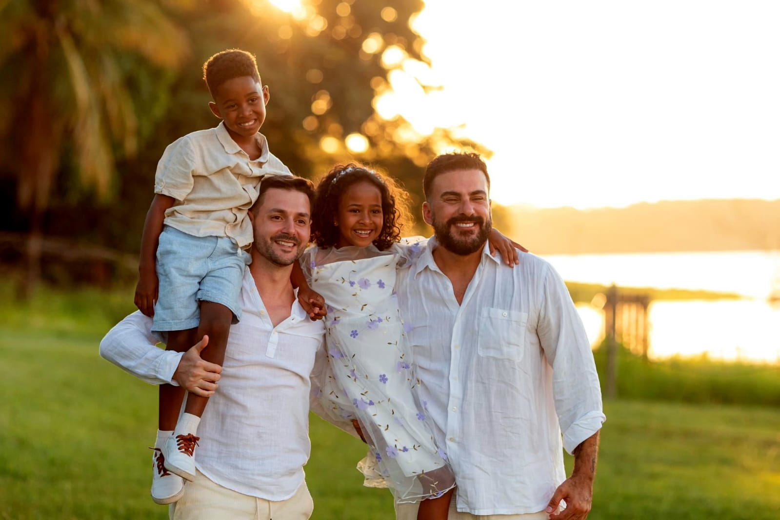 [ParaTodosVerem: Em um cenário ao ar livre, durante o pôr do sol, dois homens e duas crianças sorriem e caminham lado a lado em um gramado verde, com um lago ao fundo. O Squader João, carrega o seu filho Daniel nos ombros, enquanto o seu marido Antonio segura nos braços a Laura. Todos estão vestindo roupas leves e claras. A cena transmite afeto, união e alegria, representando uma família diversa, amorosa e cheia de conexão.]